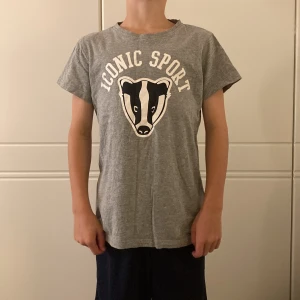 Grå t-shirt Iconic Sport med tryck - Grå t-shirt från Iconic Sport med coolt tryck av en grävling och texten 'Iconic Sport' på bröstet. Klassisk passform, rund hals och korta ärmar. Skön och mjuk i bomull, perfekt till vardags. Aldrig använd.