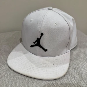 Vit Jordan keps med svart logga - Snygg vit keps från Jordan med den klassiska svarta Jumpman-loggan broderad framtill. Kepsen har rak skärm och justerbar snapback baktill för perfekt passform. Tillverkad i bomull för en skön känsla och stilren look.