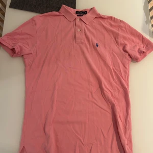 Rosa pikétröja från Ralph Lauren - Säljer en klassisk rosa pikétröja från Polo Ralph Lauren i storlek M. Tröjan har korta ärmar, krage med två knappar och den ikoniska blå broderade loggan på bröstet. Tillverkad i bomull med en skön och luftig känsla.