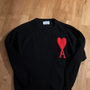 S Svart stickad tröja från Ami Paris - Svart stickad tröja från Ami Paris med rund halsringning och ikonisk röd hjärtlogga med bokstaven A på bröstet. Tröjan har långa ärmar och ribbade muddar för en clean och stilren look.
