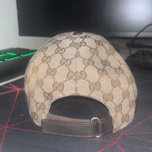 Gucci keps med GG-mönster och läder (äkta) - Snygg beige keps från Gucci med klassiskt GG-monogram över hela kepsen. Har justerbar rem i brunt läder baktill och en grön-röd rand på sidan. Materialet är bomullscanvas med detaljer i läder. Perfekt för dig som gillar exklusiva accessoarer.