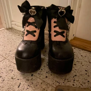 Svarta platåboots med rosetter (Jirai Kei) - Jirai kei svarta boots med hög platåsula och rosa detaljer. Klackhöjd ca 10cm. Skorna har rosa snörningspanel, svarta snören och stora svarta rosetter med glittrande hjärtformade broscher. Materialet ser ut som syntet eller skinn. Perfekta för dig som vill sticka ut med en söt men edgy stil.