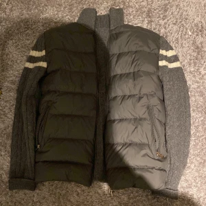 Moncler grå puffer cardigan jacka - Säljer en unik grå puffer cardigan jacka från Moncler med stickade ärmar och två vita ränder på varje arm. Jackan har quiltad framsida med dragkedjor på fickorna och hög krage. Materialmix av stickad ull och puffer-paneler ger en cool kontrast.