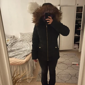 Svart pufferjacka med fuskpäls från KappAhl - Svart pufferjacka från KappAhl med snygg fuskpäls på huvan och guldfärgade detaljer vid fickorna. Jackan har quiltad design, dragkedja framtill och är perfekt för kalla dagar. Klassisk modell som håller dig varm och stilren hela vintern. Svart/Marinblå