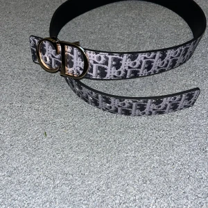 Fett Bälte - Snyggt bälte f Dmed klassiskt monogrammönster i svart och grått. Bältet har en bred design och en stor, guldig spänne med CD-logga framtill. Perfekt accessoar för att lyfta vilken outfit som helst.