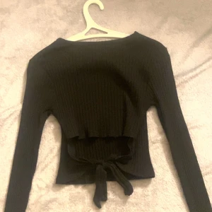 Svart ribbad långärmad topp med knyt - Svart långärmad topp från Ginatricot i ribbad bomullsmix. Modellen är croppad och har en snygg knytning framtill i midjan. Perfekt för dig som gillar stilrena och trendiga plagg med det lilla extra.