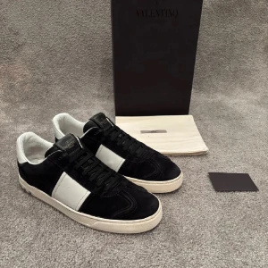 Valentino Flycrews sneakers svart/vit - Snygga Valentino Flycrews sneakers i svart mocka och vitt skinn med klassisk bred vit rand på sidan. Skorna är i UK 7 som motsvara 41-42 i EU. Skorna har svarta snören, vit sula med nitar baktill och diskret Valentino-logga på plösen. Perfekta för dig som gillar stilrena och exklusiva sneakers.