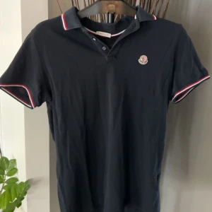 Moncler piké  - Marinblå pikétröja från Moncler med klassisk krage. Pikén har Moncler-logga på bröstet och snygga rödvita detaljer på krage och ärmslut. Tillverkad i bomull för en skön och stilren känsla.