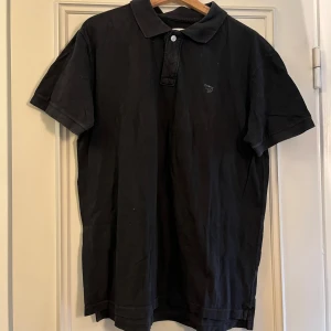 Svart pikétröja från XDYE XL - Klassisk svart pikétröja från XDYE i storlek XL. Tröjan har korta ärmar, krage och knäppning framtill. Tillverkad i mjuk bomull och har en liten broderad logga på bröstet. Perfekt för en clean och stilren look.