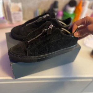 Svarta mockasneakers med dragkedja - Snygga svarta sneakers från New Balance i mocka med stilren dragkedja framtill istället för klassisk snörning. Skorna har platt sula och rund tå, samt detaljer i svart läder vid hälen. Perfekta för dig som gillar en clean och modern look. Plus de finns en skada på ett par av skorna jag har tagit bild på den men när man har på sig skorna så ser man inte skadan den är inte heller så synlig