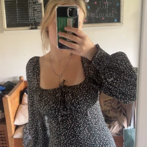 Svart blommig kortklänning med puffärm - Supersöt svart kortklänning med småblommigt mönster i vitt och rosa. Klänningen har långa puffärmar, fyrkantig urringning och en söt knytning framtill. Figurnära passform som framhäver midjan och ger en romantisk vibe.