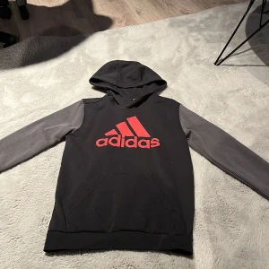 Svart och grå Adidas hoodie med röd logga - Svart hoodie från Adidas med grå ärmar och stor röd Adidas-logga på bröstet. Tröjan har huva och känguruficka framtill. Perfekt för dig som gillar sportig och avslappnad stil.