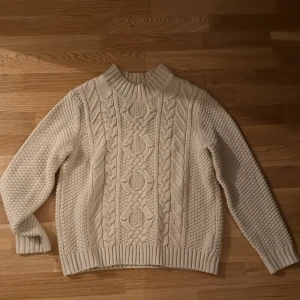 Beige stickad tröja med flätmönster - Säljer en beige stickad tröja med grovt flätmönster framtill och strukturstickade ärmar och rygg. Tröjan har hög ribbad krage och muddar vid ärmslut och nederkant. Perfekt för kyliga dagar och supermysig att ha på sig. Passar xs också