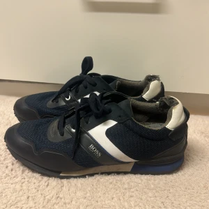 Svarta sneakers från BOSS - Snygga svarta sneakers från BOSS med vita och blå detaljer. Skorna har snörning, rund tå och är tillverkade i en kombination av textil och syntetmaterial. Perfekta för en sportig och stilren look.