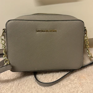 Beige axelväska från Michael Kors - Snygg beige axelväska från Michael Kors med guldfärgade kedjedetaljer och justerbar rem. Väskan har en stilren, fyrkantig form och är tillverkad i ett tåligt skinnmaterial med diskret struktur. Perfekt för dig som vill ha en klassisk och trendig accessoar.