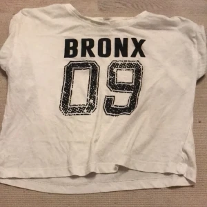 Vit t-shirt Bronx 09 Red & Blue - Vit t-shirt från Red & Blue med tryck 'BRONX 09' i svart på framsidan . Klassisk passform och korta ärmar, tillverkad i mjuk bomull. Perfekt för en sportig och avslappnad stil.