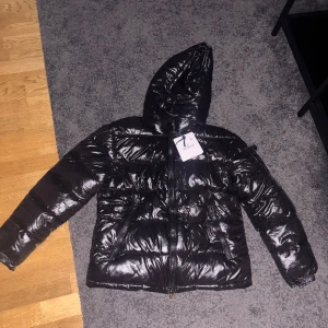 Svart Moncler jacka - Säljer en svart, glansig pufferjacka  Moncler med huva och dragkedja framtill. Jackan har Moncler-logga på ärmen, två fickor med dragkedja och quiltad design. Perfekt för kalla dagar och riktigt snygg streetstil. Storlek M.