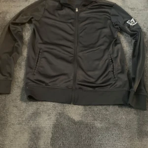 Svart EA7 ziptröja från Emporio Armani - Svart ziptröja från Emporio Armani EA7 med logga på ärmen och dragkedja framtill. Tröjan har ribbade muddar, två fickor och är tillverkad i ett mjukt, sportigt polyestermaterial. Perfekt för en clean och sportig look.