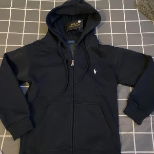Mörkblå hoodie från Polo Ralph Lauren - Mörkblå hoodie från Polo Ralph Lauren med dragkedja, huva med snörning och klassisk logga broderad på bröstet. Tröjan har ribbade muddar och känguruficka framtill. Perfekt för en chill och stilren look.
