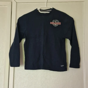 Mörkblå långärmad tröja från Morxs - Mörkblå långärmad tröja från Morxs med tryck på bröstet där det står 'COLLEGE' och 'Los Angeles'. Tröjan har rund halsringning och är gjord i mjuk bomull. Perfekt för en avslappnad. Under höger arm hänger det trådar, men som syns på sista bilden är det inget hål utan bara trådar.