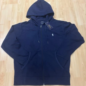 Marinblå hoodie från Polo Ralph Lauren - Snygg marinblå hoodie från Polo Ralph Lauren med dragkedja och huva. Tröjan har klassisk logga broderad på bröstet, fickor framtill och justerbar snörning i huvan. Tillverkad i mjuk bomull för en skön känsla.