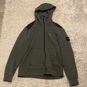 Stone Island grön hoodie med huva - Snygg grönhoodie från Stone Island med svarta axelpartier och klassisk logga på ärmen. Tröjan har hel dragkedja, huva och två fickor framtill. Perfekt för dig som gillar streetwear och vill ha något stilrent men ändå unikt.
