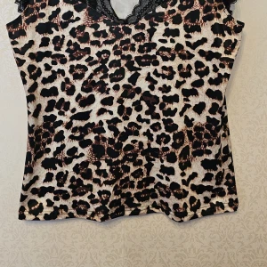 Leopardmönstrad topp med spetsdetaljer - Säljer en snygg leopardmönstrad topp med svarta spetsdetaljer vid ärmar och v-ringning. Toppen har en figurnära passform och är ärmlös med spets som ger en extra touch. Perfekt för dig som vill sticka ut med ett djurmönster och coola detaljer.