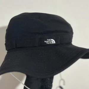 Matcha den franska stilen med denna bekväma fiske haten med snöre🇫🇷 - Svart bucket hat från The North Face med broderad logga framtill. Hatten har bred brätte, ventilationshål på sidorna och justerbar dragsko under hakan. Tillverkad i lätt och slitstark nylonblandning, perfekt för uteliv och streetstyle.