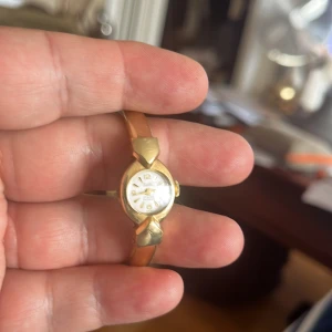 Guldfärgad Weitz damklocka - Snygg guldfärgad damklocka från Weitz med oval urtavla i vitt och tydliga siffror. Armbandet är stelt och har en elegant, smal design med dekorativa detaljer vid urtavlan. Klockan är schweizisk och har 17 rubiner enligt urtavlan. Perfekt för dig som gillar klassisk stil.