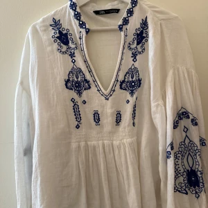 Vit och blå broderad blus från Zara - Supersöt vit blus från Zara med blå broderade detaljer och v-ringad hals. Långärmad med bohemisk vibe och snygga mönster på både bröst och ärmar. Tillverkad i lätt linne, perfekt för varma dagar.