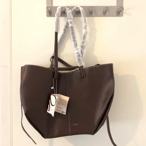 Brun axelväska från Polène i skinn - Snygg brun axelväska från Polène i slätt skinn med vita kontrastsömmar. Väskan har en rymlig form och kommer med en matchande mindre väska med dragkedja. Perfekt för dig som gillar stilrena och praktiska accessoarer.