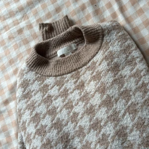Beige och vit stickad polotröja H&M - Supermysig stickad polotröja från H&M i beige och vitt med klassiskt rutigt mönster. Tröjan har ribbade muddar, hög krage och är långärmad. Perfekt för lager på lager och chill höst- och vinterstil.