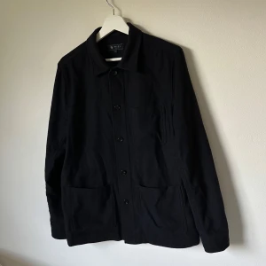 Svart overshirt från Riley i ullmix - Stilren svart overshirt från Riley i storlek 48. Jackan har klassisk krage, knäppning framtill och stora fickor. Tillverkad i en mjuk ullmix (60% ull, 35% polyester, 5% polyamid) som ger en clean och modern look. 2I4