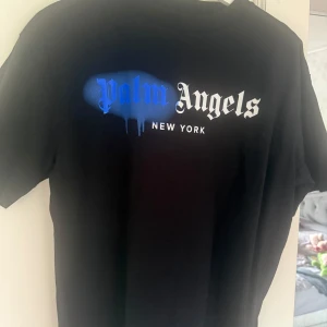Svart Palm Angels t-shirt med blå print - Svart t-shirt från Palm Angels med korta ärmar och rund hals. Framsidan har ett stort Palm Angels-tryck i blått och vitt med coolt drip-effekt och texten 'New York'. Materialet är mjuk bomull, perfekt för streetwear. Märkesetikett i nacken och originaltagg medföljer. Inköpt via Miinto innan sommaren, använd 2ggr, tvättad en gång. 