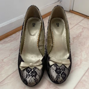 Svarta spetsklädda pumps med rosett - Eleganta pumps med svart spets och blommigt mönster. Skorna har en söt rosett framtill och smal klack. Perfekta för dig som vill sticka ut med en klassisk men unik look.