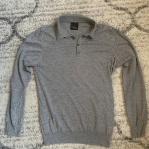 Grå pikétröja från Oscar Jacobson - Snygg grå pikétröja från Oscar Jacobson i mjuk bomull. Tröjan har klassisk krage, tre knappar framtill och långa ärmar. Perfekt för en clean och stilren look. Passar dig som gillar enkel men sofistikerad stil.