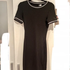 Svart ribbad klänning från Divided - Svart ribbad kortklänning från Divided med vita ränder på krage och ärmslut. Klänningen har en tight passform, korta ärmar och rund halsringning. Perfekt för dig som gillar en sportig och stilren look.