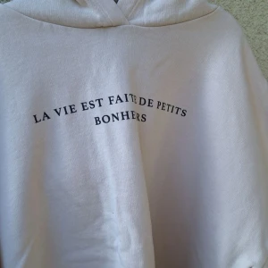 Beige hoodie med fransk text - Beige hoodie med svart tryck framtill där det står 'La vie est faite de petits bonheurs'. Tröjan har huva, långa ärmar och dragsko nertill. Perfekt för en chill och avslappnad stil.