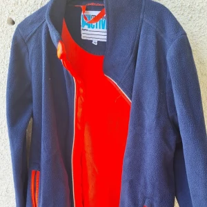 Blå fleecejacka med röda detaljer - Mörkblå fleecejacka från Name It Active med röda detaljer på dragkedja och fickor. Jackan har hög krage och är helfodrad i rött, perfekt för kyliga dagar. Snygg och sportig look som passar till många outfits.