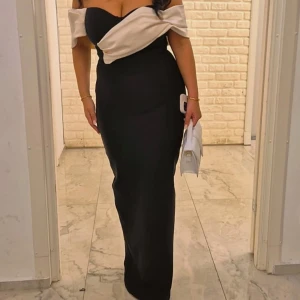 Svart långklänning - Elegant långklänning i svart med beige offshoulder-detalj upptill. Klänningen har figurnära passform och en snygg axelbandslös design. Perfekt för fester. Använd endast två gånger och är i storlek S.