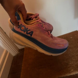 Hoka Clifton 9 - Säljer ett par snygga Hoka sneakers i rosa med blå detaljer och vit sula. Skorna har snörning och är tillverkade i ett lätt syntetmaterial med mesh för bra andningsförmåga. Perfekta för dig som gillar att springa eller vill ha riktigt bekväma skor till vardagen.