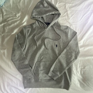 Grå Ralph Lauren hoodie - Snygg grå hoodie från Polo Ralph Lauren med klassisk broderad logga på bröstet. Tröjan har huva med snörning, känguruficka och ribbade muddar. Perfekt för en chill och stilren look. Tillverkad i mjuk bomullsmix för extra komfort.