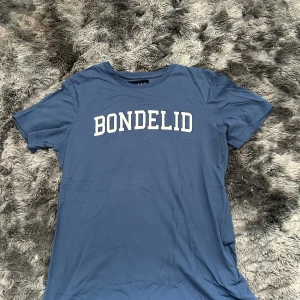 Blå t-shirt från Bondelid, storlek S - Snygg blå t-shirt från Bondelid med vit text framtill. Klassisk rund halsringning och korta ärmar. Tillverkad i mjuk bomull som känns skön mot huden. Perfekt för en avslappnad och stilren look.
