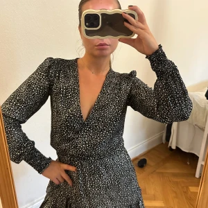 Svart kortklänning med vita prickar - Supersnygg svart kortklänning med vita hjärtan och djup v-ringning. Klänningen har långa ärmar med resår vid handlederna och volang nertill. Perfekt för dig som gillar en lekfull och trendig stil.