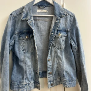 Ljusblå jeansjacka från Lee - Säljer en klassisk ljusblå jeansjacka från Lee med snygga sömmar och silverfärgade knappar. Jackan har två bröstfickor med lock och knapp, samt en rak passform och krage. Perfekt för dig som gillar en tidlös och avslappnad stil.