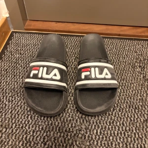 Svarta Fila badsandaler - Svarta sandaler från Fila med klassisk logga i vitt och rött på remmen. Snygg och enkel design med platt sula och öppen tå, perfekta för stranden eller poolen. Tillverkade i syntetmaterial för en lätt och bekväm känsla.