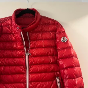 Röd dunjacka från Moncler - Snygg röd dunjacka från Moncler med klassisk quiltad design och logga på ärmen. Jackan har dragkedjor i vitt och silver, hög krage och två fickor med dragkedja. Perfekt för dig som vill sticka ut med en ikonisk och trendig vinterjacka.