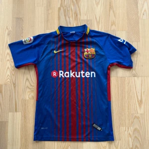 FC Barcelona Messi 10 Nike fotbollströja - Säljer en officiell FC Barcelona fotbollströja från Nike med Messi 10-tryck på ryggen. Tröjan är blå och röd med smala vertikala ränder, gula detaljer och klubbmärke på bröstet. Tillverkad i lätt och ventilerande Dri-FIT-material. Perfekt för dig som älskar Barca och Messi. Små svarta prickar som inte syns så mycket , finns på bilden 