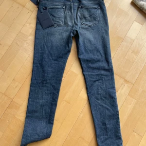 Mörkblå jeans från Jacob Cohen - Snygga mörkblå jeans från Jacob Cohen med klassisk femficksdesign och diskret slitning framtill. Jeansen har raka ben och normal passform, tillverkade i mjukt bomullstyg med en hint av stretch för extra komfort. Perfekta för dig som gillar stilrena och tidlösa jeans.