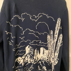 Mörkblå stickad tröja med ökenmotiv från Vans - Säljer en mörkblå stickad tröja från Vans med ett unikt ökenmotiv i vitt. Tröjan har långa ärmar och ribbade muddar. Perfekt för dig som gillar streetstyle och vill sticka ut med coolt mönster.